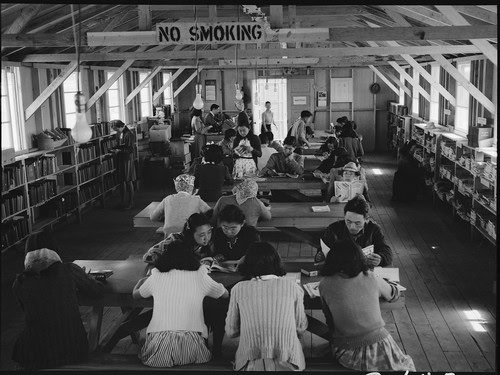 Group of people reading and studying in a library with bookshelves and a 'No Smoking' sign.