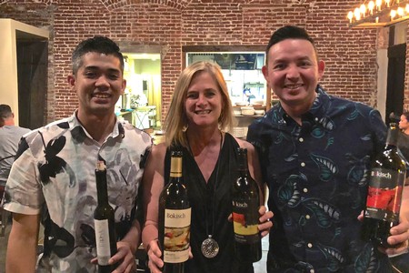 Three people holding wine bottles at Lodi Winegrape Commission event, smiling in a brick-walled venue.