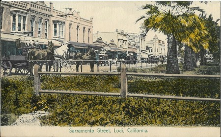 Vintage photo of Sacramento Street in Lodi, California, showing old buildings, horse-drawn carriages, and trees.
