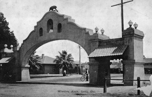 Black and white photo of Mission Arch in Louis Cal with palm trees and a cyclist nearby.