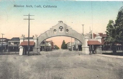 Historic Mission Arch in Iodi, California, with a wide street and surrounding buildings, under a clear sky.