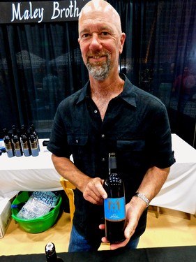 Man holding a wine bottle at Lodi Winegrape Commission event, smiling, with a black curtain backdrop.