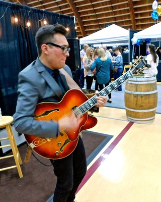 Young man playing guitar at Lodi Winegrape Commission event with attendees in background.