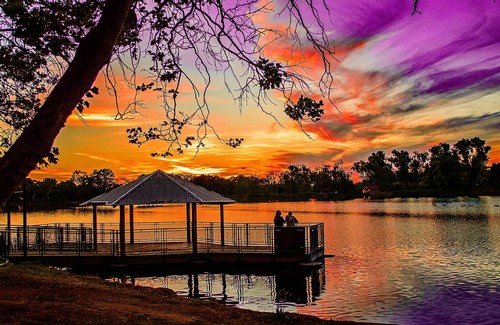 Scenic sunset over a lake with a dock and pavilion, vibrant purple and orange sky, trees reflected in water, peaceful outdoor.