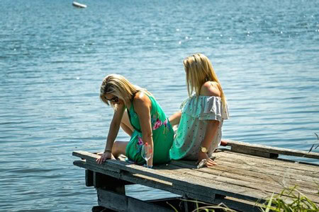 Two young girls sitting on a wooden dock by the water, enjoying a sunny day outdoors.