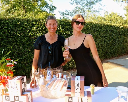 Two women at a wine tasting event outdoors, smiling, with wine glasses and promotional materials on the table.