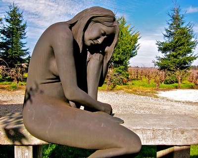 Bronze sculpture of a seated woman in a vineyard with trees and blue sky in the background.