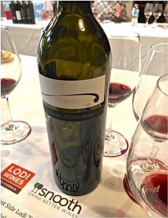 A wine bottle from Lodi Winegrape Commission with glasses of red wine on a table.