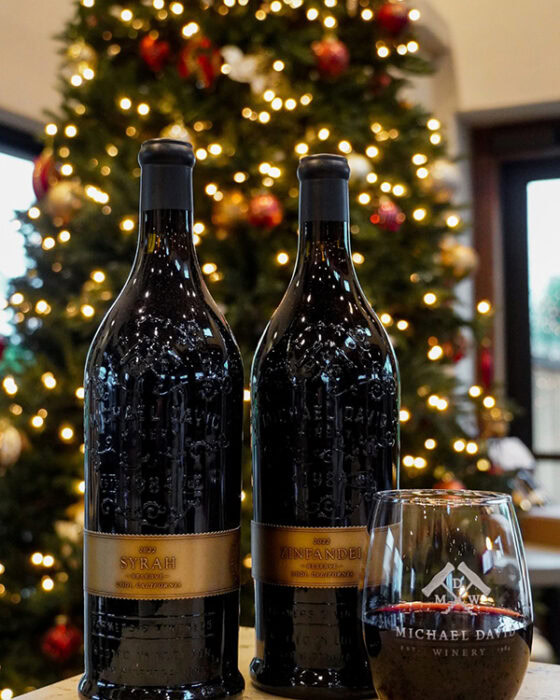 Two bottles of wine and a glass in front of a decorated Christmas tree in a winery setting.