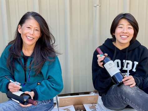 Two women smiling at a wine tasting event, holding wine bottles, with a beige background.