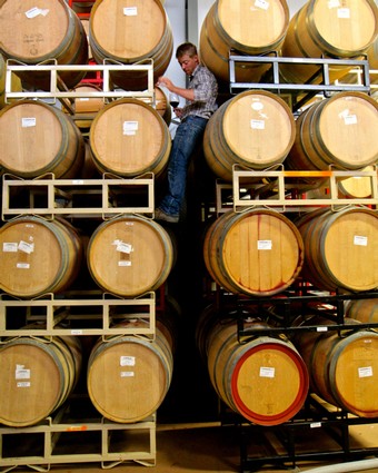 Person inspecting wine barrels in a winery storage facility with stacked barrels around.