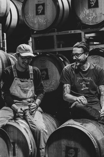 Two people enjoying wine tasting at a winery with barrels in the background, smiling and engaging in conversation.