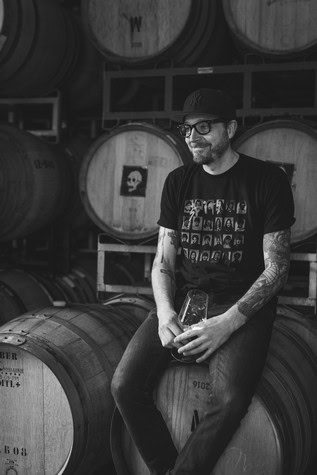 Man with glasses and tattoos enjoying wine at Lodi wine cellar with barrels in background.