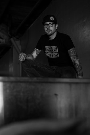 Black and white portrait of a man with glasses and tattoos, wearing a Lodi Winegrape Commission T-shirt, in a winery setting.