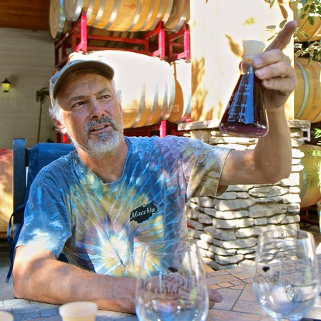 Man examining a wine sample in a vineyard setting with wine barrels in the background.