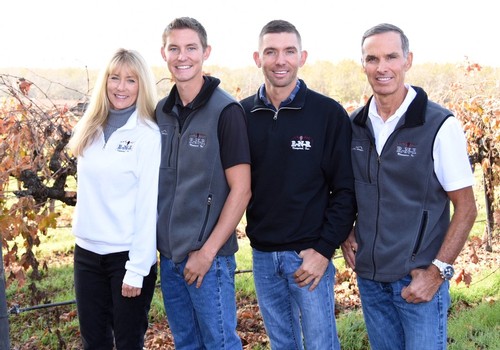 Group of four people standing in a vineyard, smiling, with grapevines in the background.