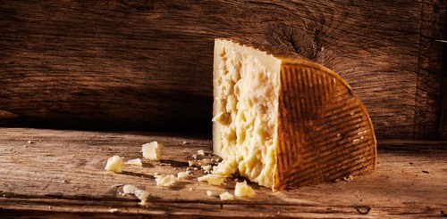 A wedge of cheese with a textured rind on a rustic wooden table, with cheese crumbs nearby.