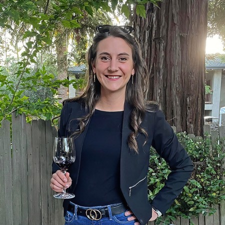 Woman smiling with a glass of red wine outdoors, standing near a large tree and greenery.