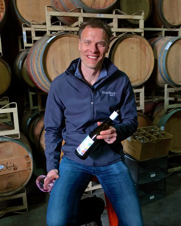 Man holding wine bottle and glass in wine cellar with barrels in background.