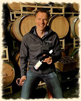 Man holding a wine bottle and glass at a wine cellar with barrels in the background.