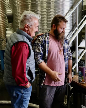 Two men inspecting wine in a winery, one older and one younger, with wine tanks in the background.