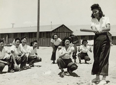 Women playing baseball outdoors in a rural area with a barn and utility poles in the background.