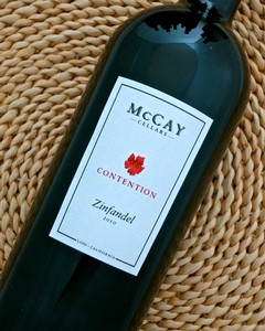 Close-up of a McCay Cellars Zinfandel wine bottle on a woven surface, showcasing the label and branding.