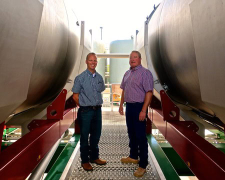 Two men in checkered shirts standing between large wine tanks at a winery.