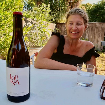 Woman smiling at outdoor table with wine bottle and glass, lush garden background, promoting Lodi winegrapes.