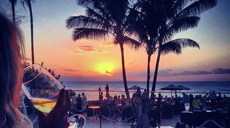 Sunset over the ocean with palm trees and people enjoying drinks at a beachside event.