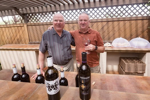 Two men at a wine tasting event with bottles of wine on a wooden table.