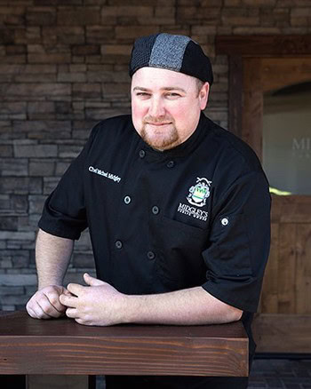 Portrait of a chef in black uniform with Lodi Winegrape Commission logo, smiling, in a rustic setting.