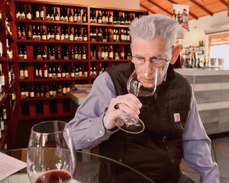 Senior man tasting wine at Lodi Winegrape Commission tasting room with wine bottles in background.
