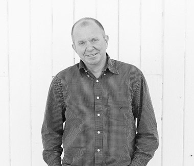 Man in checkered shirt smiling in front of a white wooden wall.