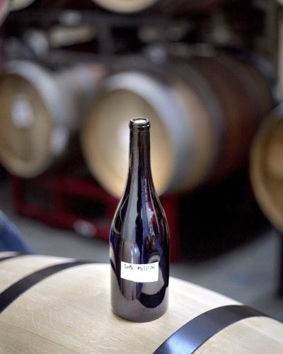 A dark glass wine bottle with a handwritten label in a wine cellar or barrel room setting.
