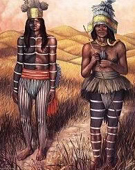 Two African women in traditional clothing and body paint standing in a field with a landscape background.