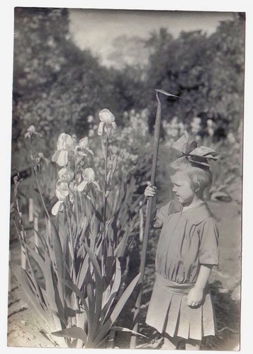 Young girl in vintage dress holding a kite string in a garden with tall flowers and trees in the background.