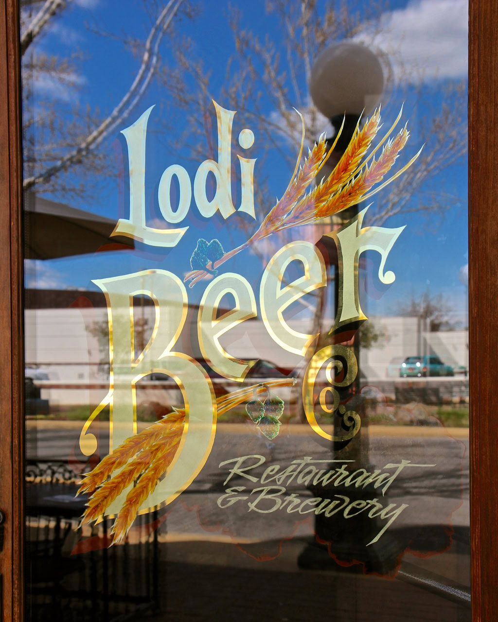 Lodi Winegrape Commission sign on glass door with wheat graphic and restaurant info, reflecting outdoor scene.
