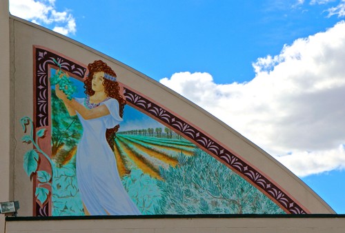 Colorful mural of a woman in a white dress holding grapes, with vineyards in the background, under a blue sky.