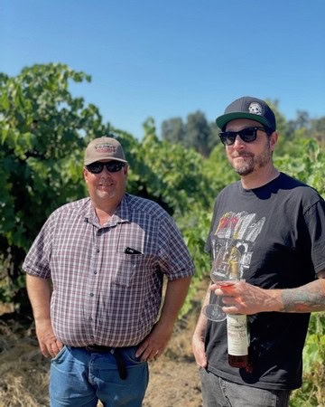 Two men in vineyard, one holding a wine bottle, enjoying a sunny day at Lodi Winegrape Commission.