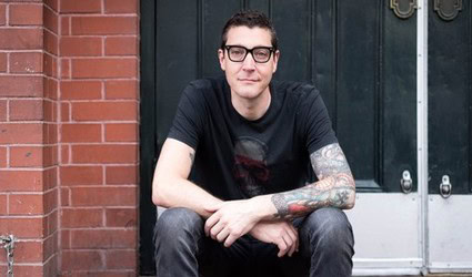 Young man with glasses and tattoos sitting outside near a brick wall and green door.