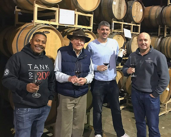 Group of four men tasting wine in a winery with wine barrels in the background.