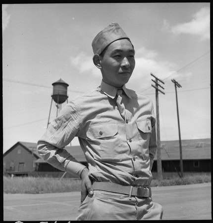 Black and white photo of a woman in military uniform standing outdoors with buildings and power lines in the background.