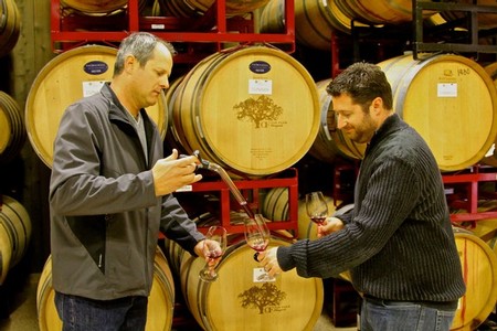 Two men tasting wine in a cellar with wine barrels in the background.