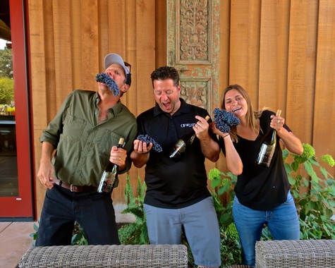 Three people celebrating with wine bottles and grapes in front of a wooden backdrop.