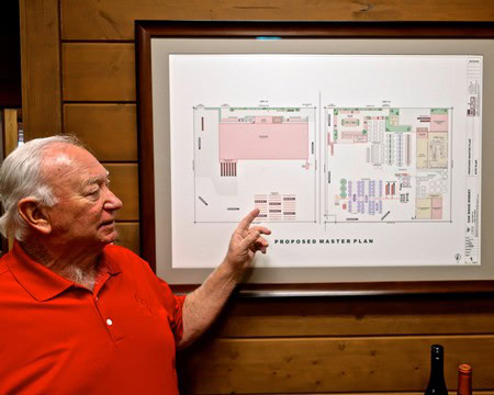 Senior man in red shirt pointing at a detailed master plan diagram on a wall.
