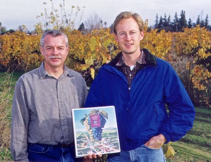 Two men standing in a vineyard holding a Lodi Winegrape Commission brochure, with grapevines in the background.