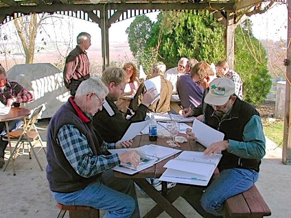Group of people at a table outdoors, reviewing documents, with vineyard and trees in the background.