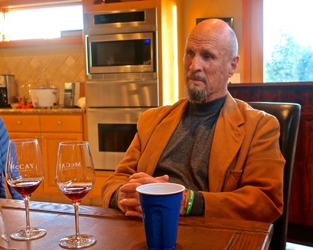 Man at a table with wine glasses and a coffee cup, in a kitchen setting, discussing wine and grape industry.