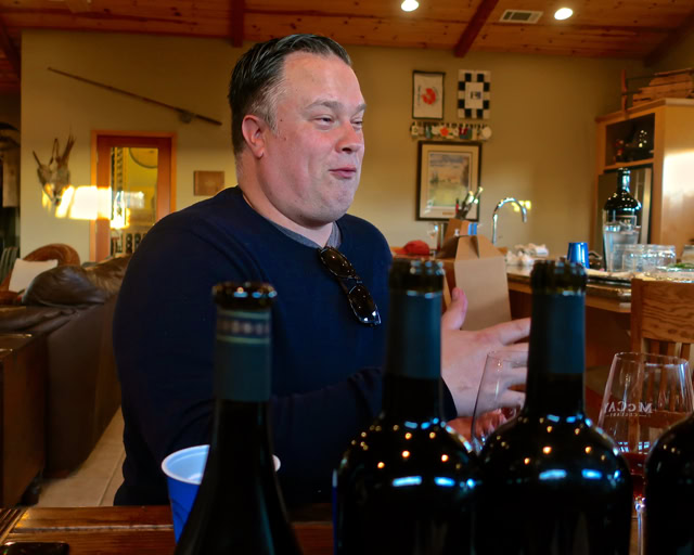 Man at a wine tasting event with bottles of wine in the foreground and a cozy indoor setting.